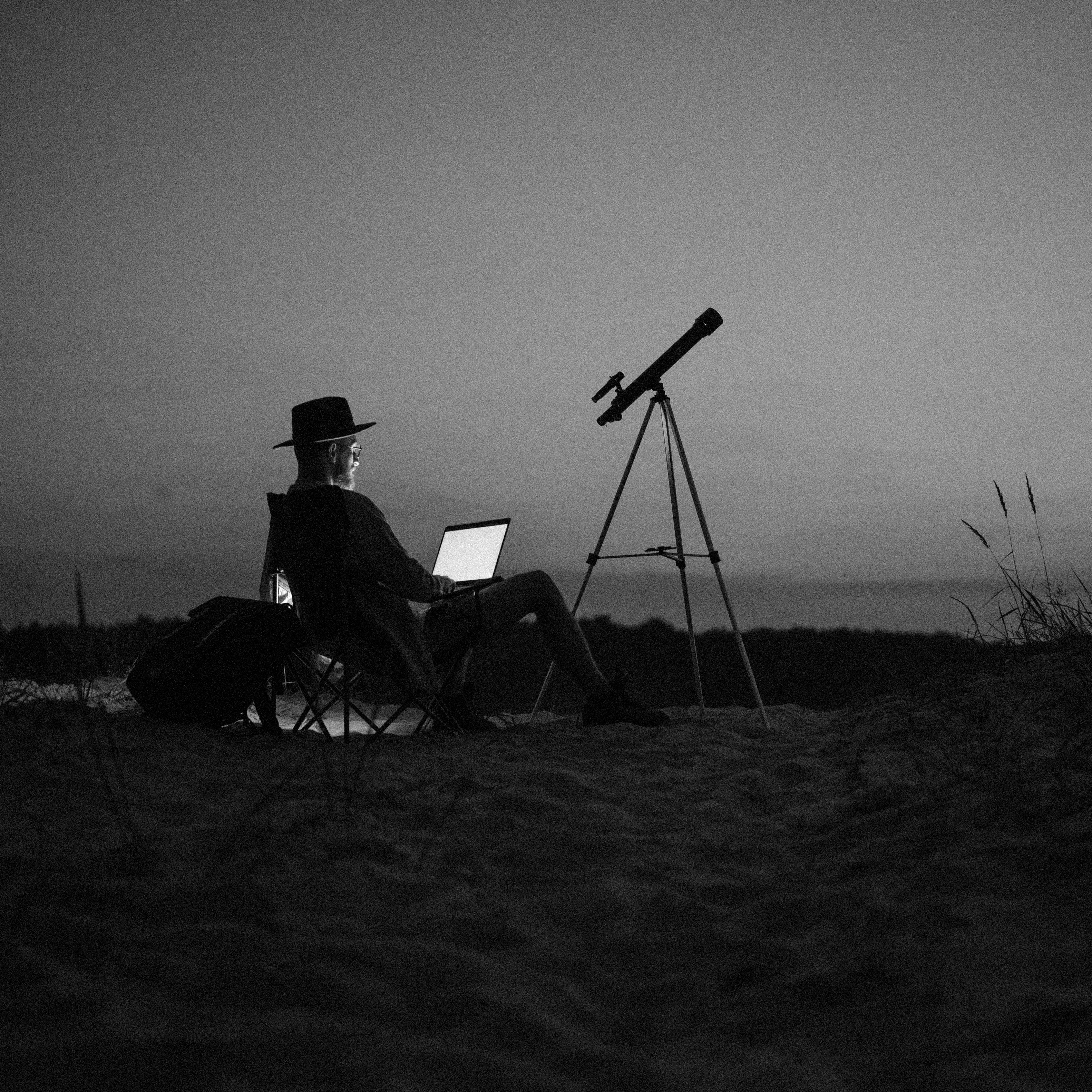 Fotografia przedstawia mężczyznę siedzącego obok teleskopu skierowanego w niebo.